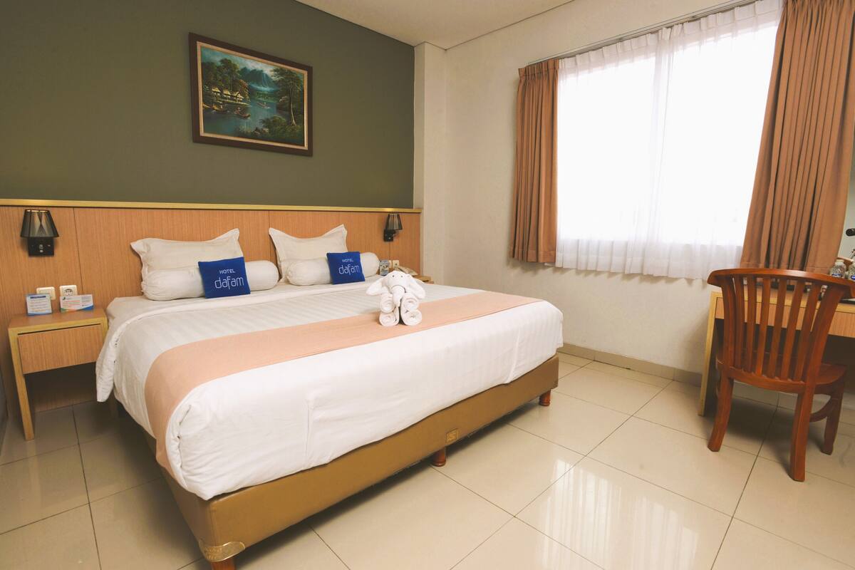 A hotel room with a large bed, a wooden headboard, a nightstand with a lamp, a desk with a chair, and a painting on the wall.