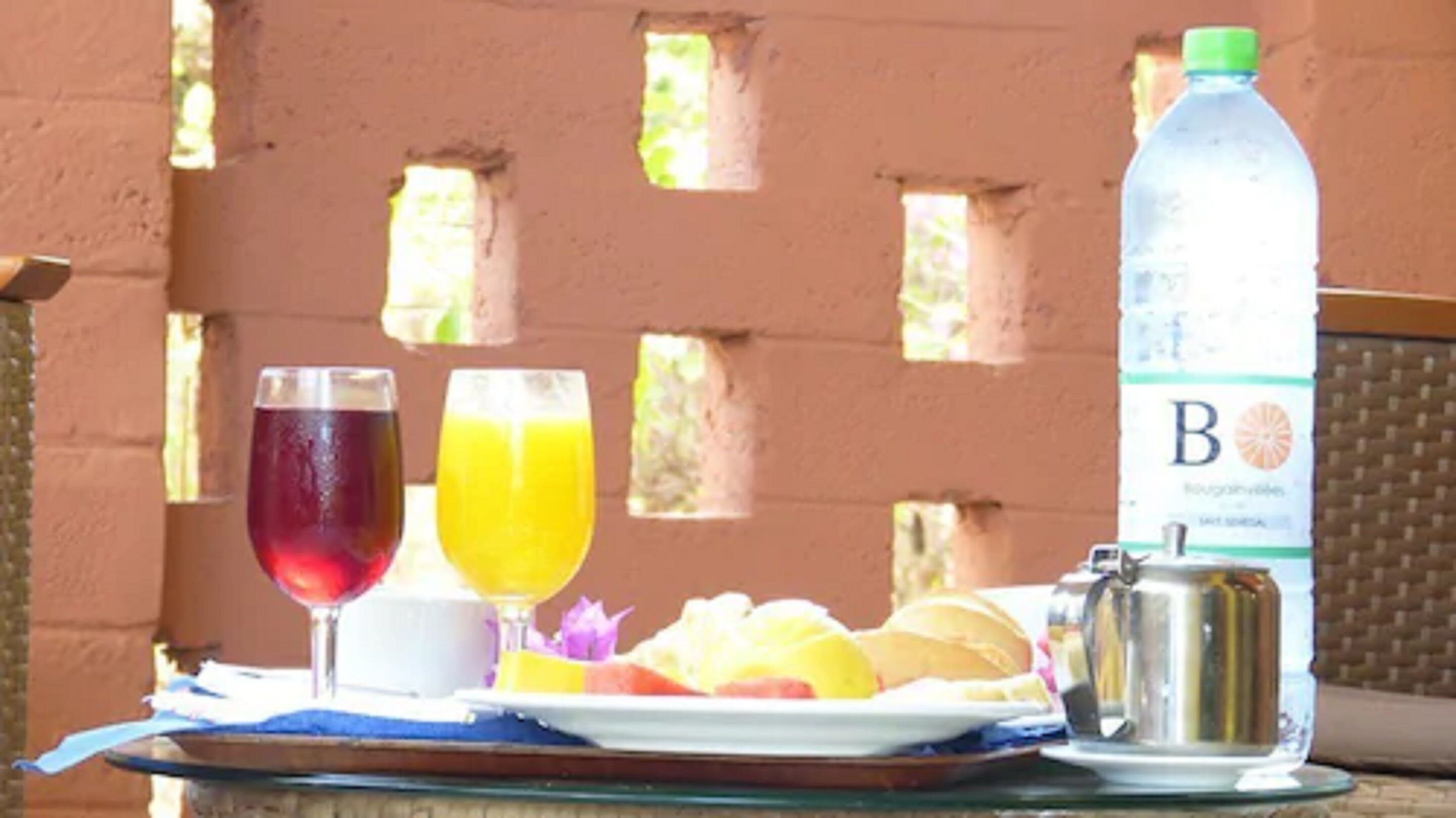 Un plateau avec un verre de boisson rouge, un verre de jus d’orange, une assiette de fruits, une bouteille d’eau et une cafetière.