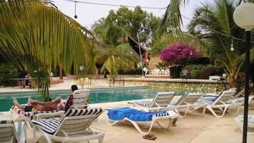Un área junto a la piscina con sillones de descanso y una piscina, rodeada de palmeras y plantas tropicales.