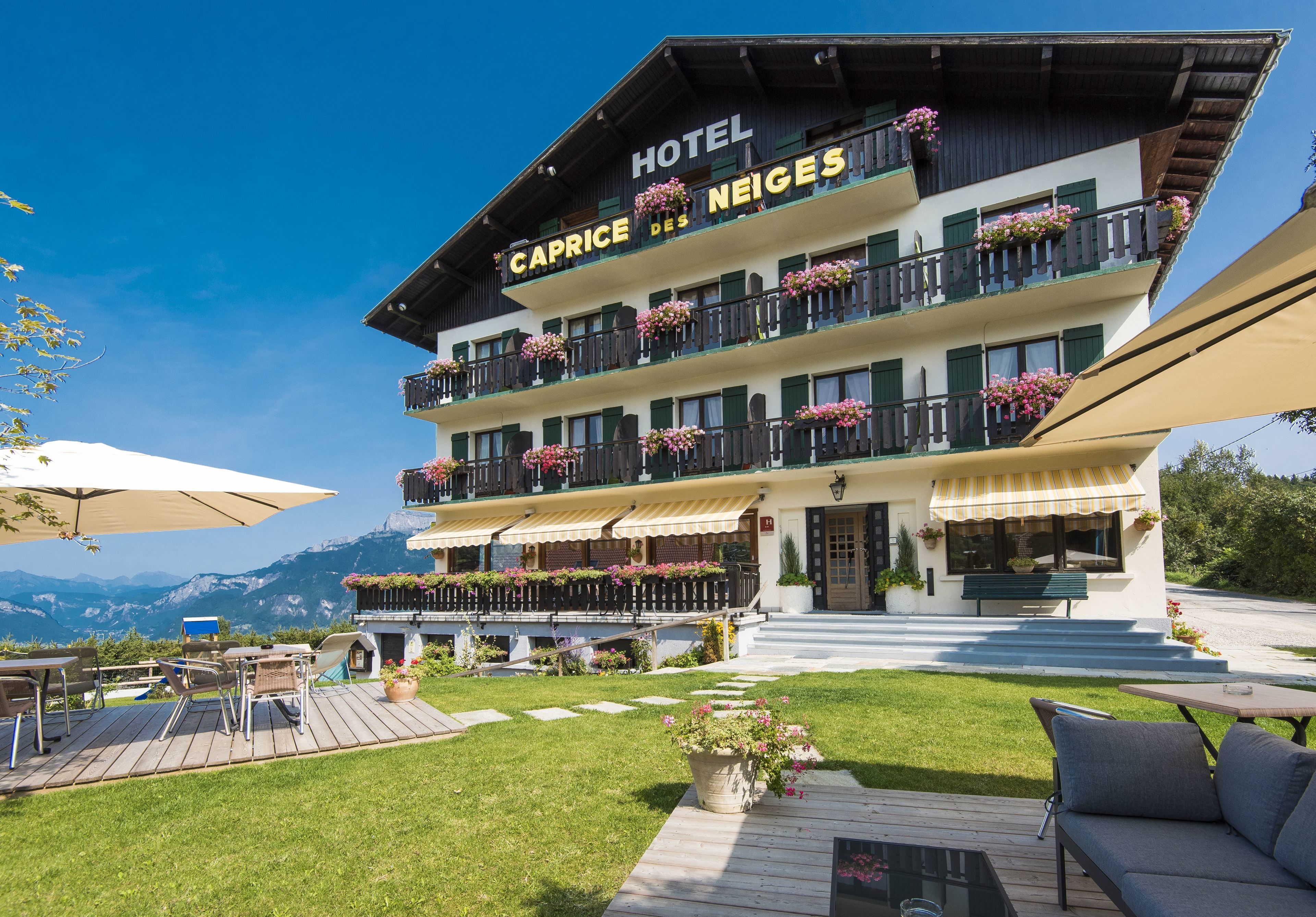 Ein Hotel mit Balkonen, die mit Blumen geschmückt sind, einer Terrasse mit Sitzgelegenheiten im Freien und Blick auf Berge.