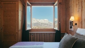 A room with a bed, a window with a view of mountains, a wooden wall, and a wall-mounted lamp. - Caprice Des Neiges (Combloux)