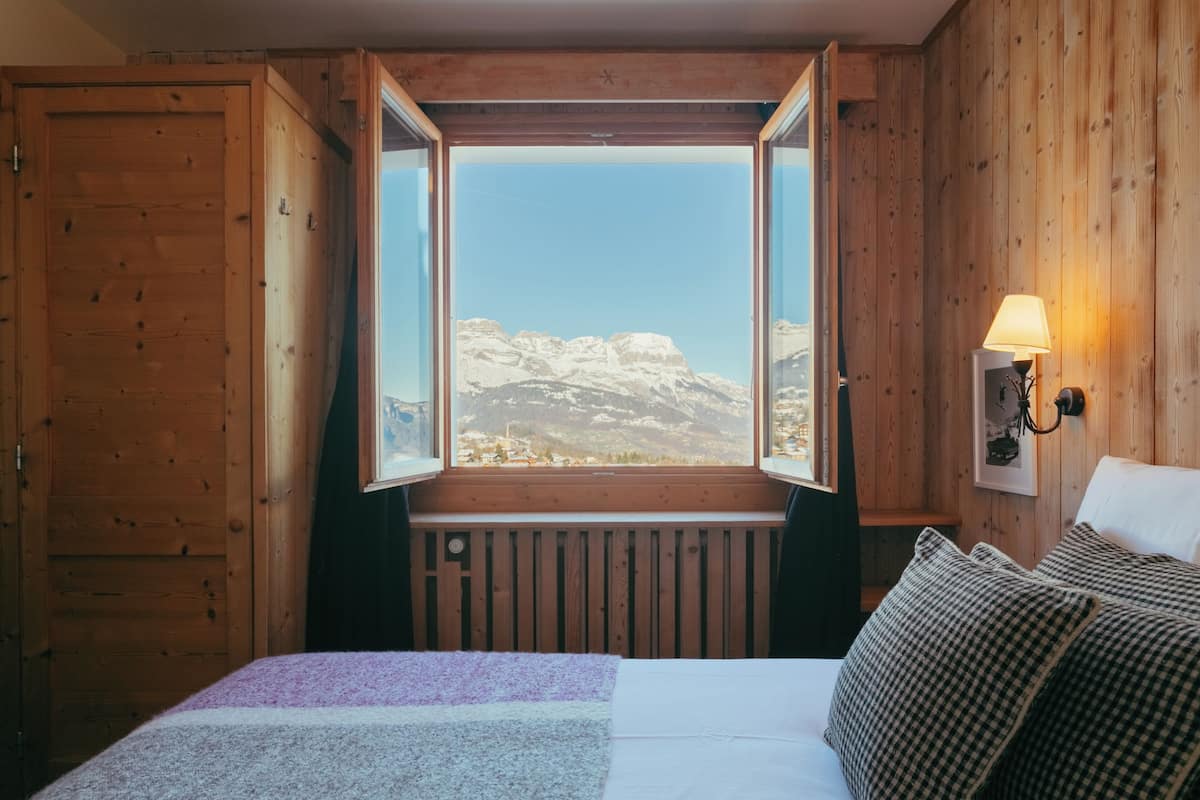 Una habitación con una cama, una ventana que ofrece vistas a montañas, una pared de madera y una lámpara fijada a la pared.