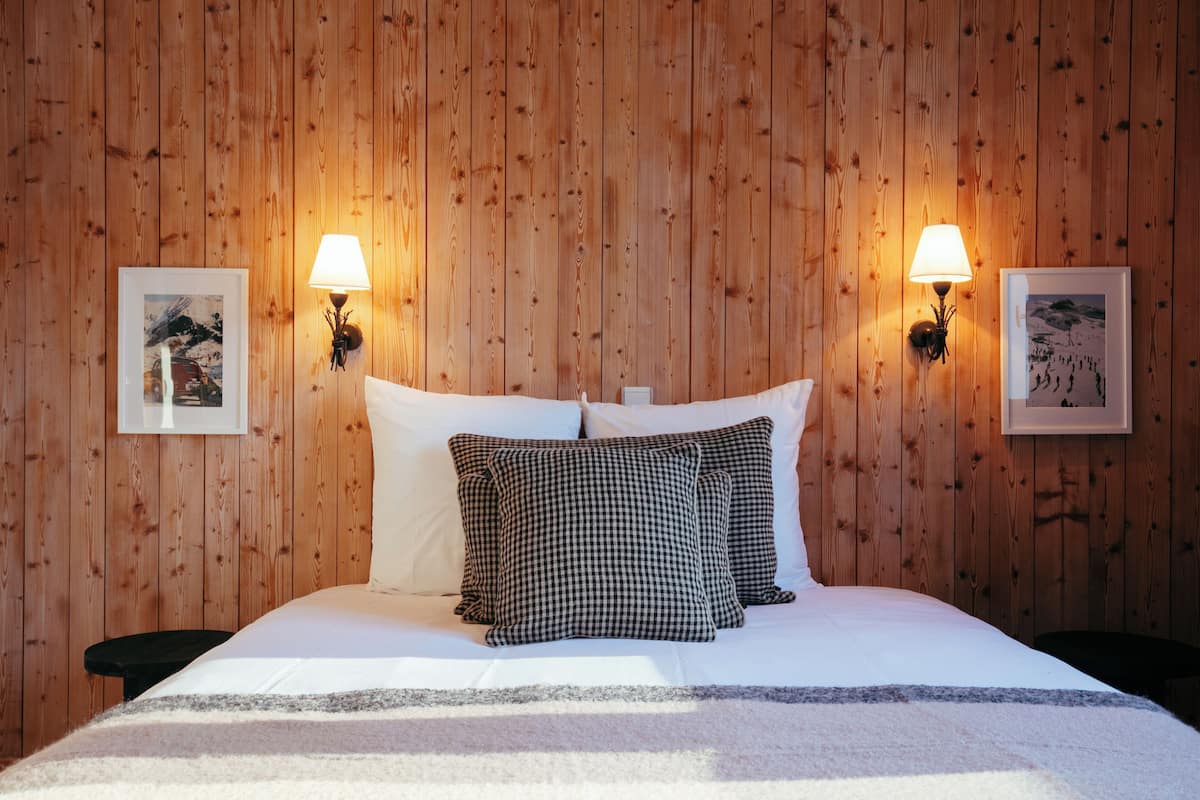 Una cama bien hecha con almohadones blancos y negros a cuadros, dos lámparas fijadas a la pared y cuadros en una pared de madera.