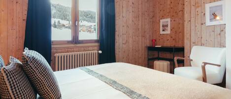 A wooden-paneled room with a bed, a chair, a radiator, and a window with curtains.