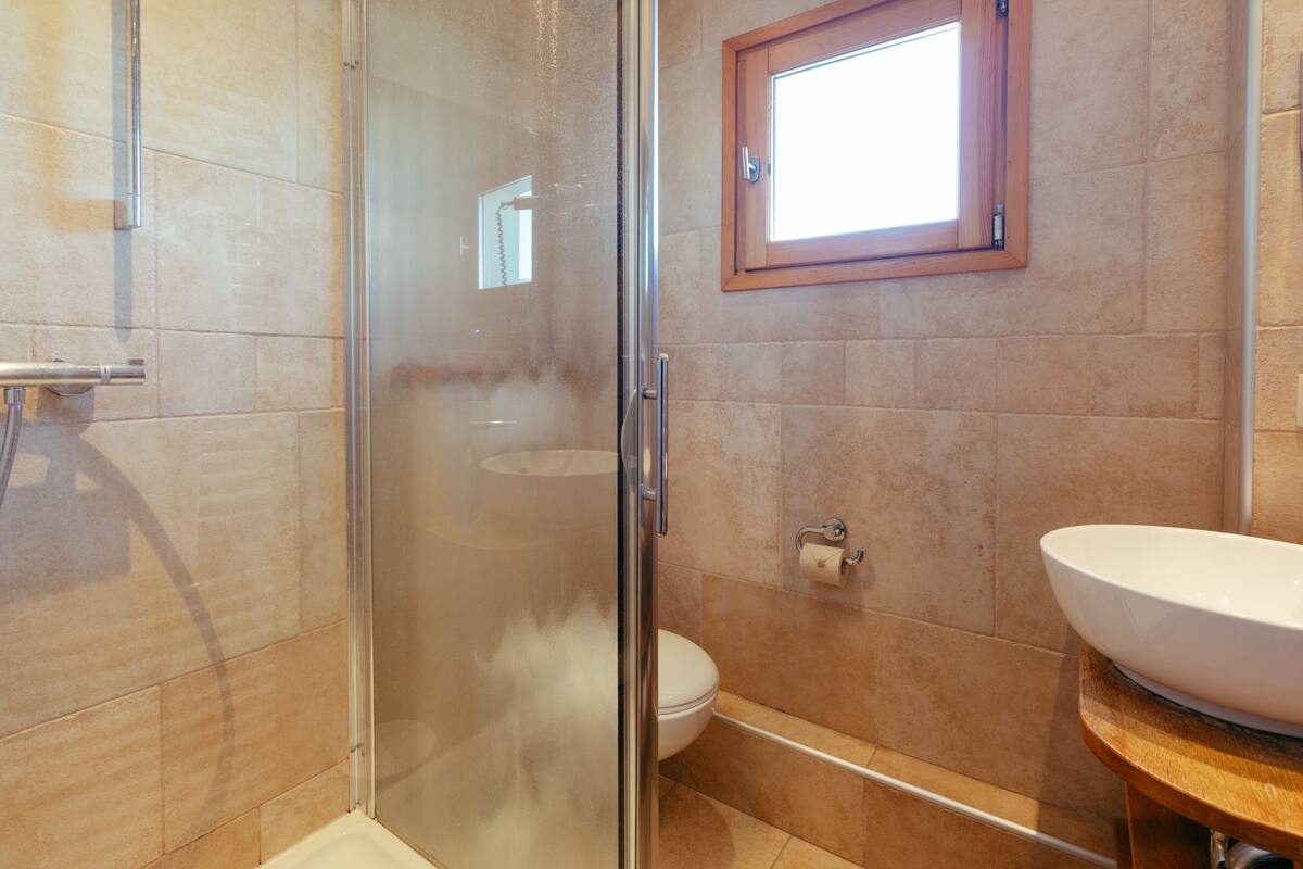 Un baño con cabina de ducha de vidrio, ventana con marco de madera, lavabo blanco y un inodoro.