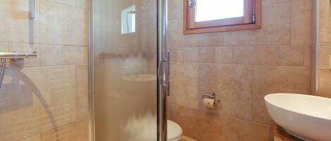 Un baño con cabina de ducha de vidrio, ventana con marco de madera, lavabo blanco y un inodoro.
