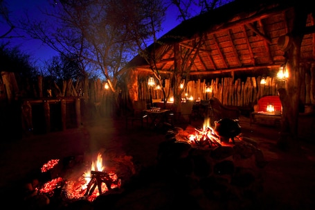 A rustic outdoor dining area with a thatched-roof structure, tables, chairs, and a central fire pit.