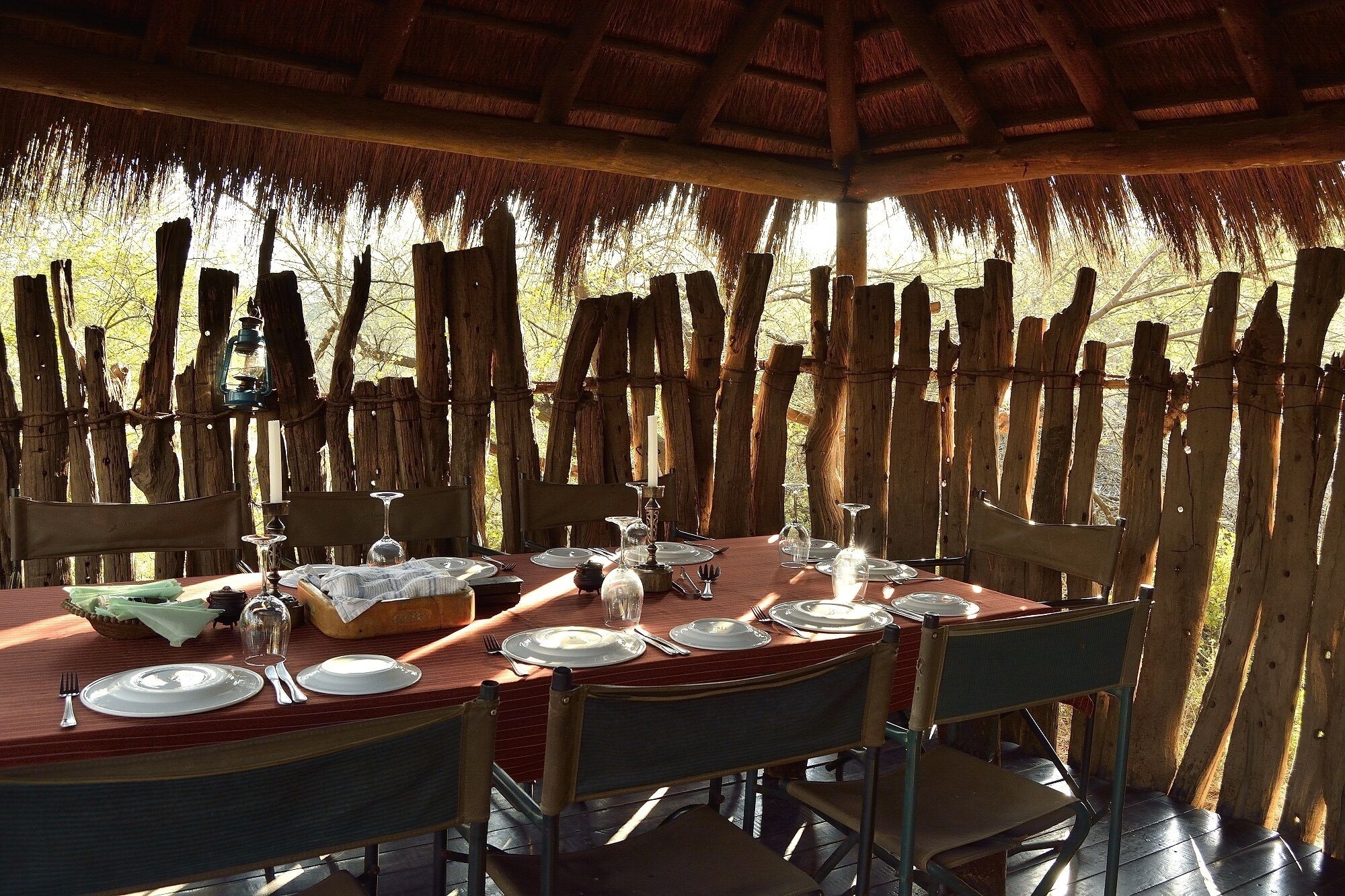 An outdoor dining area with a thatched roof, wooden chairs, and a table set for a meal.