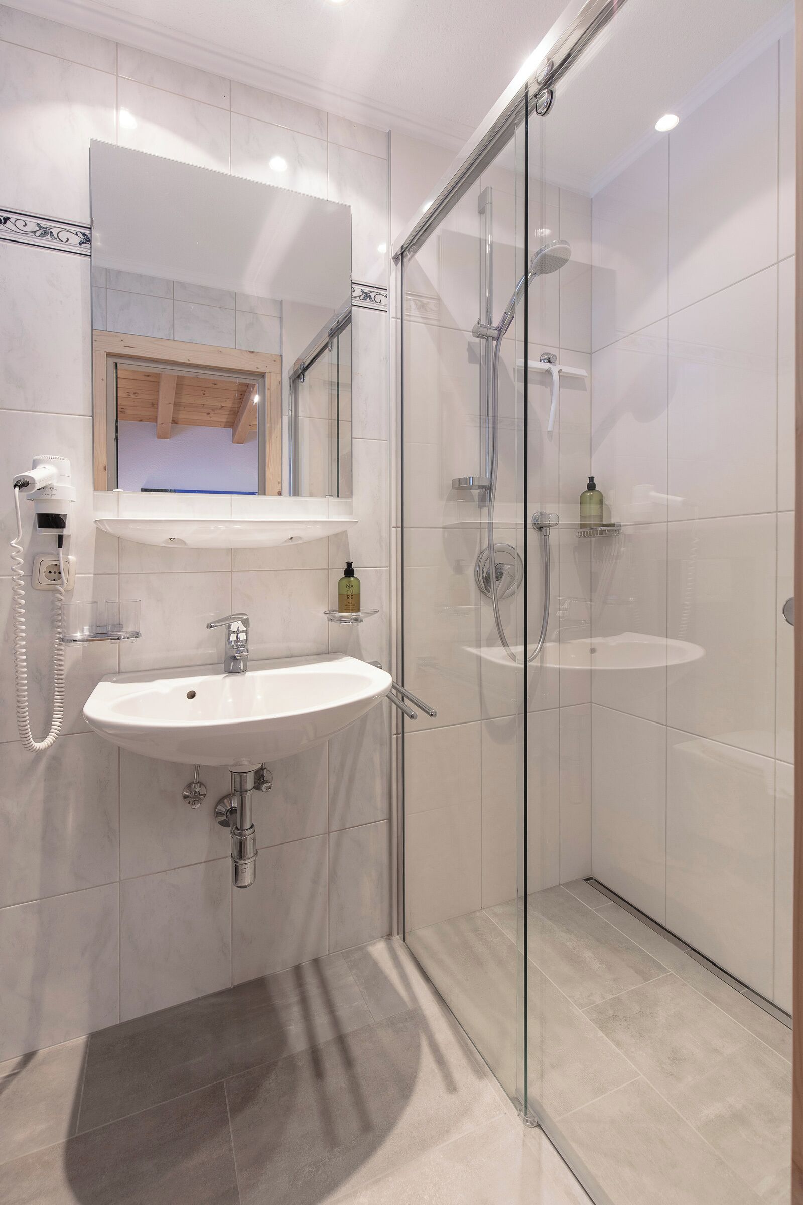 Une salle de bain moderne avec une douche à parois de verre, un lavabo blanc avec un miroir au-dessus et une petite étagère contenant des articles de toilette.