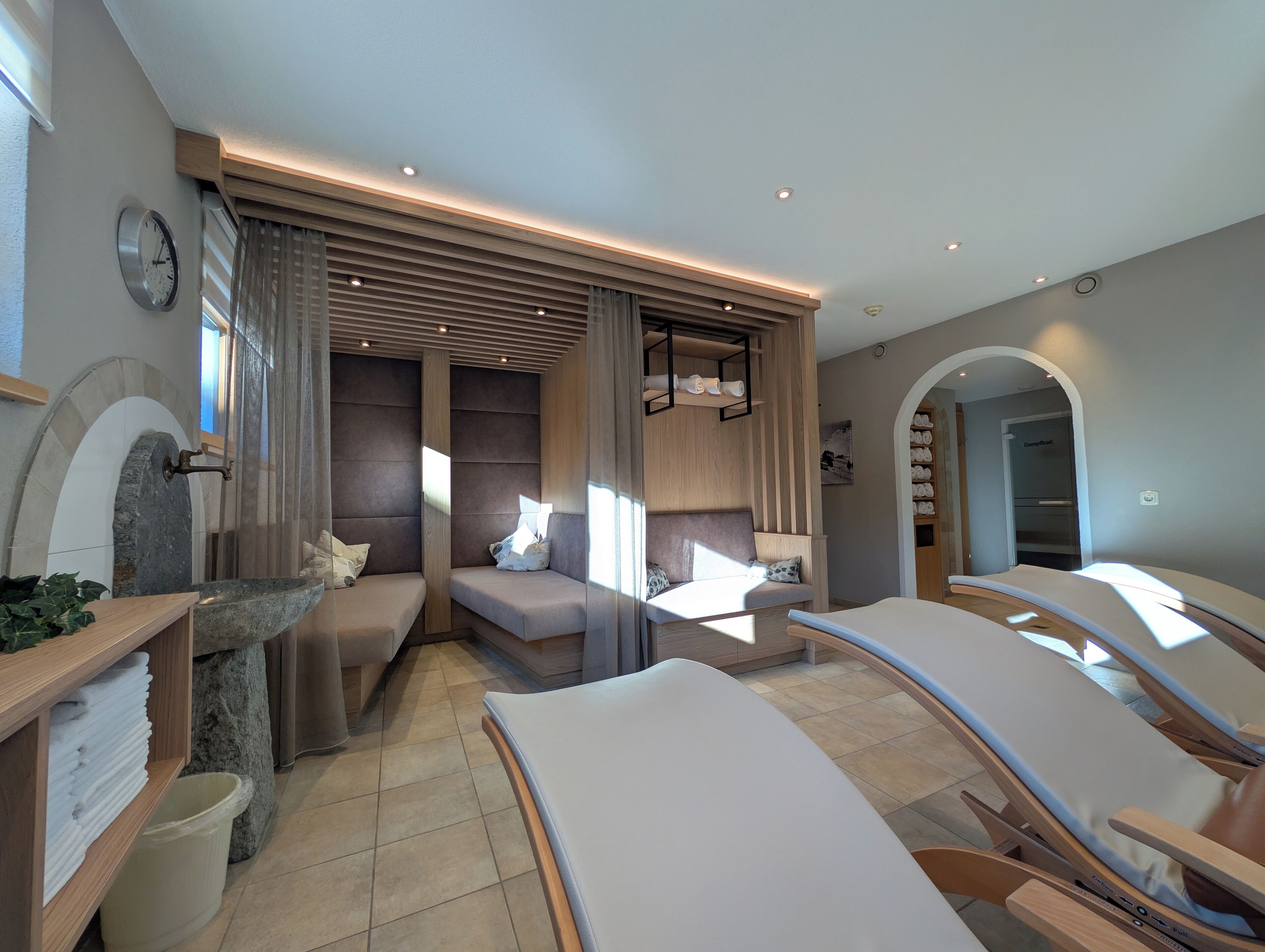 Ein moderner Spa-Raum mit einem Steingefäß, zwei Sitzbereichen mit Kissen sowie einem Entspannungsbereich mit Massageliege.