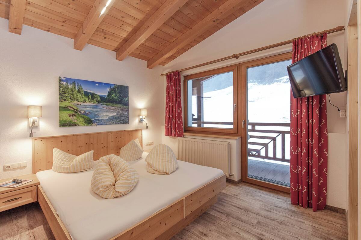 A bedroom with a wooden bed, a mounted TV, a window with curtains, and a painting of a landscape.