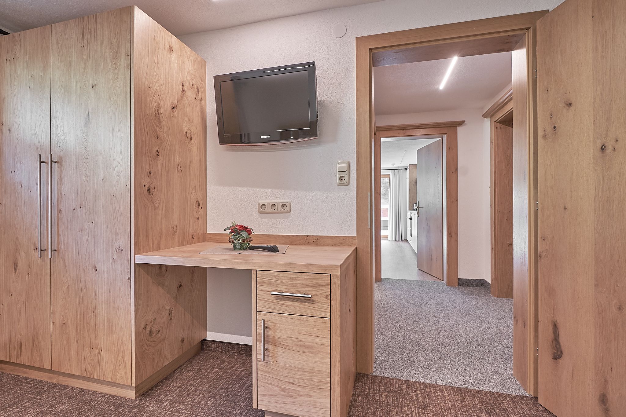 A room with a wooden desk, a flat-screen TV, and a door leading to another room.