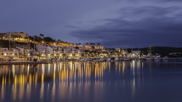 Un puerto deportivo con barcos amarrados, edificios a lo largo del muelle y aguas tranquilas que reflejan las luces.