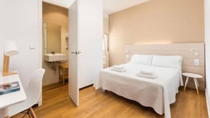 Una habitación de hotel moderna con suelo de madera, una cama con ropa blanca, un escritorio con lámpara y un baño visible a través de una puerta abierta.