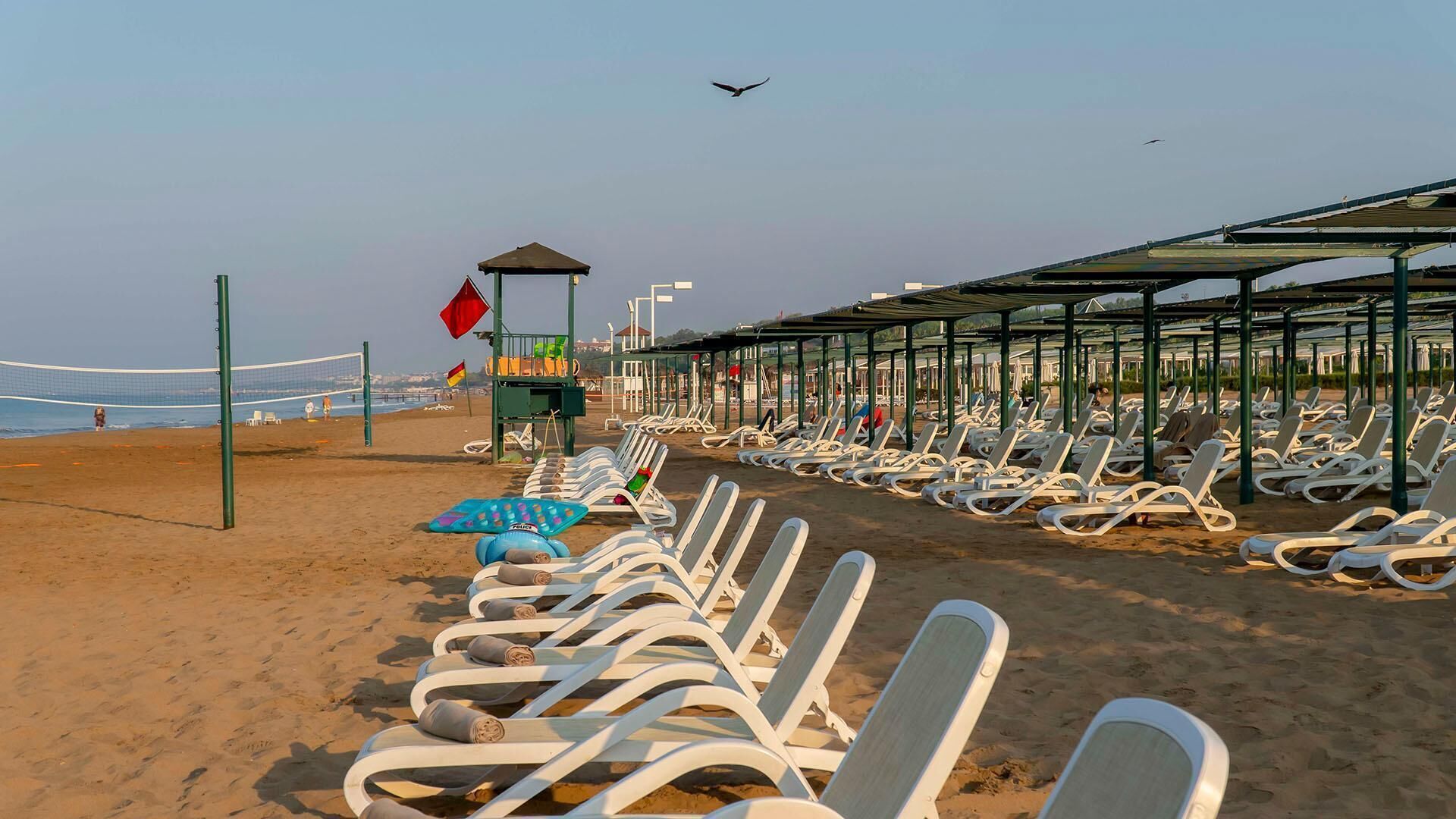 A beach with rows of white sun loungers under a covered area, a volleyball net, and a lifeguard station with flags.