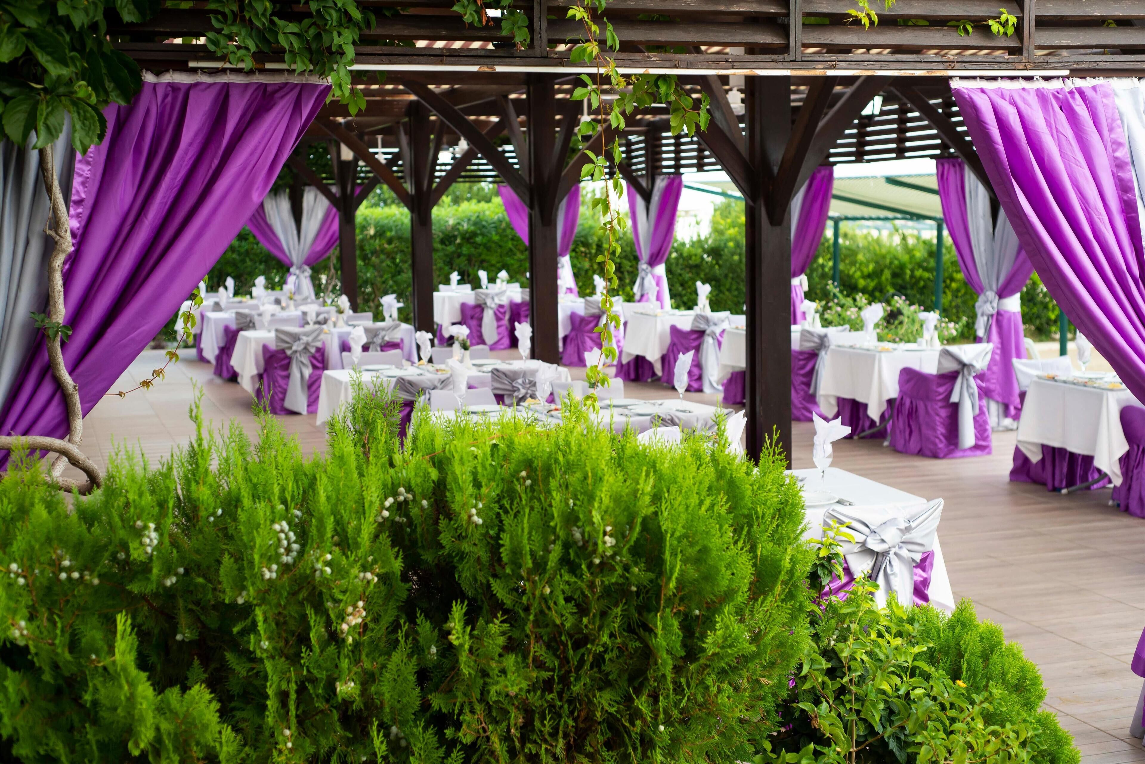 Un espacio al aire libre para eventos con mesas dispuestas para una recepción, con decoración morada y blanca.