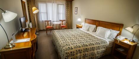 Habitación de hotel con una cama grande, un escritorio con computadora, una silla, una mesita y una ventana con cortinas.