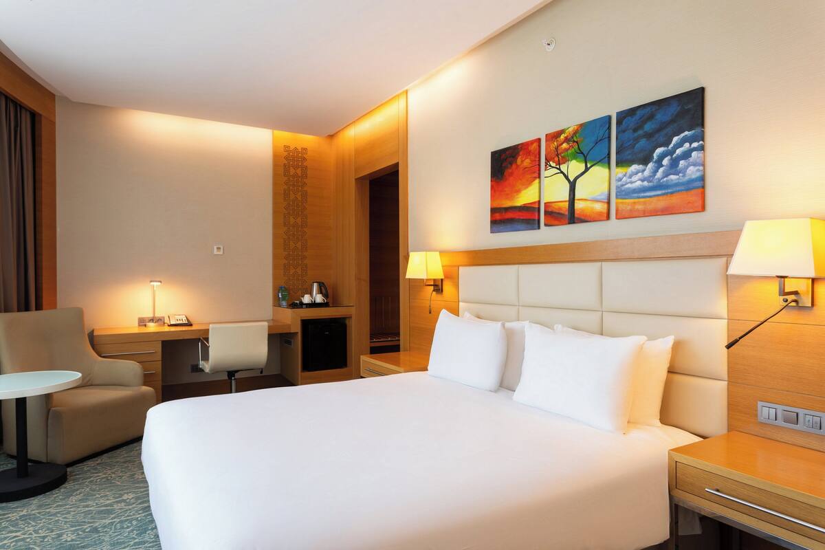 Una habitación de hotel con una cama grande, un escritorio con silla, una mesita y un cuadro en la pared.