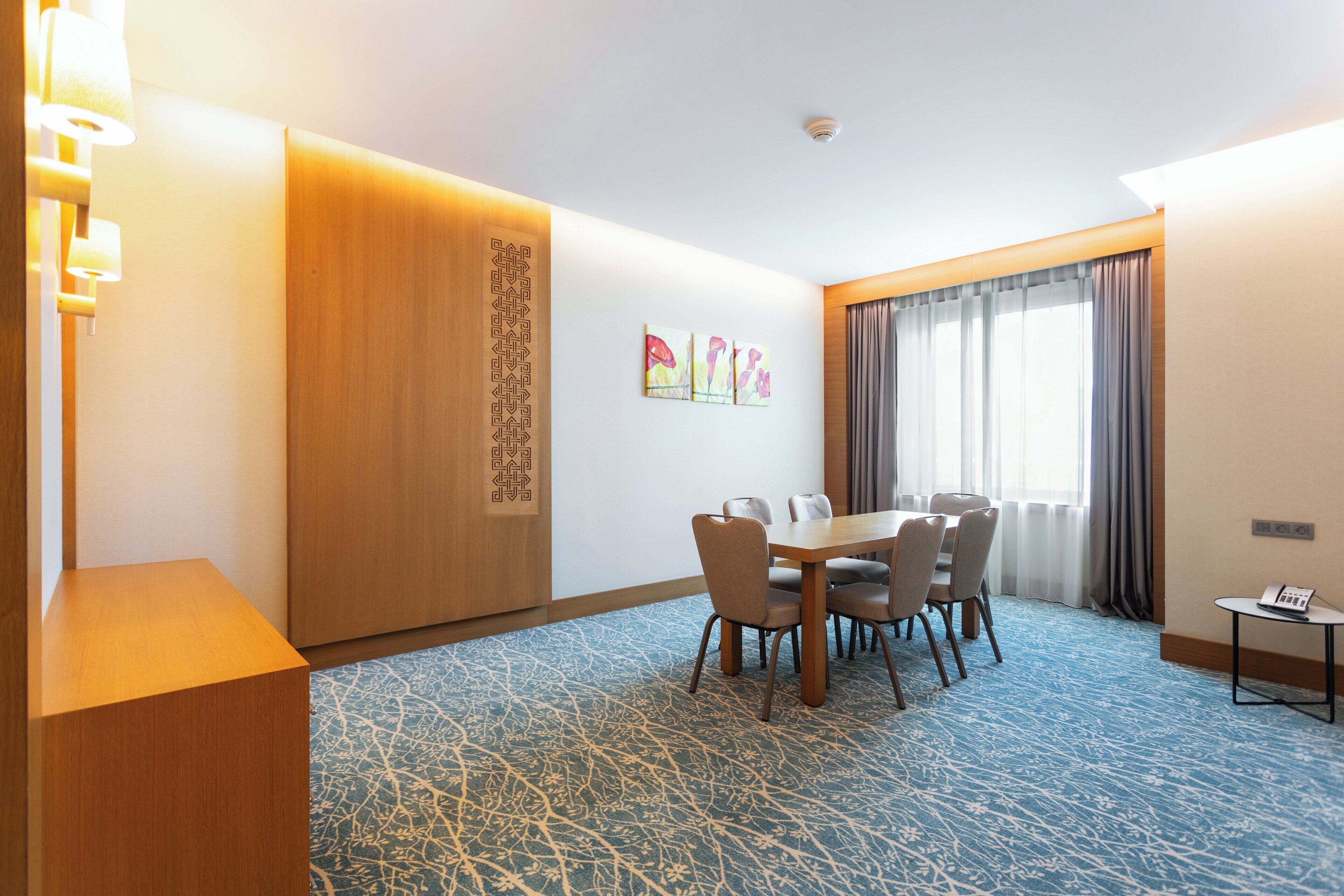 Ein Hotelzimmer mit einer Holzwand, einem Esstisch mit Stühlen, einem kleinen Tisch mit Telefon und einem Fenster mit Vorhängen.
