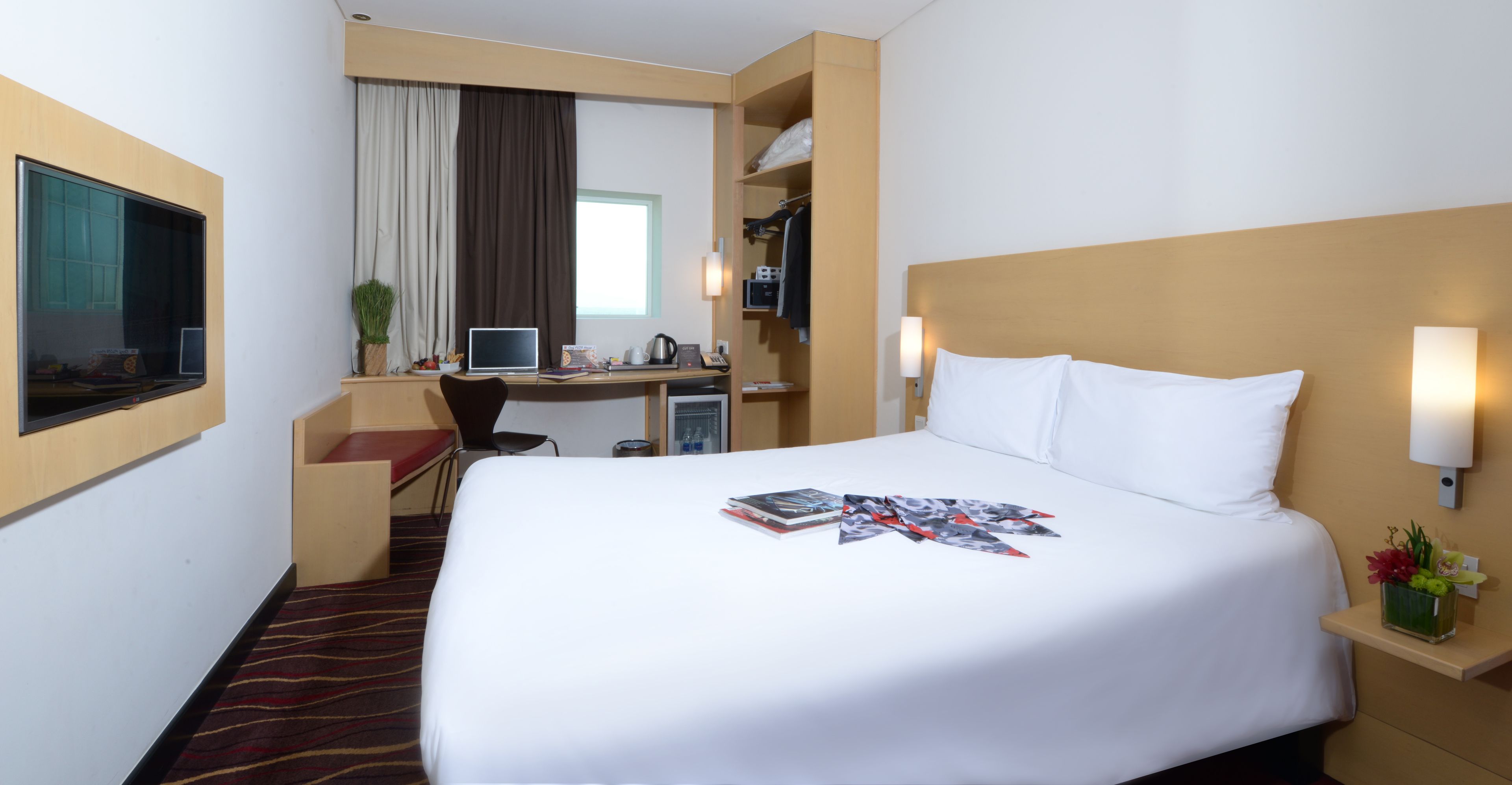 A hotel room with a large bed, a desk with a computer, a chair, and a small table with a vase of flowers.