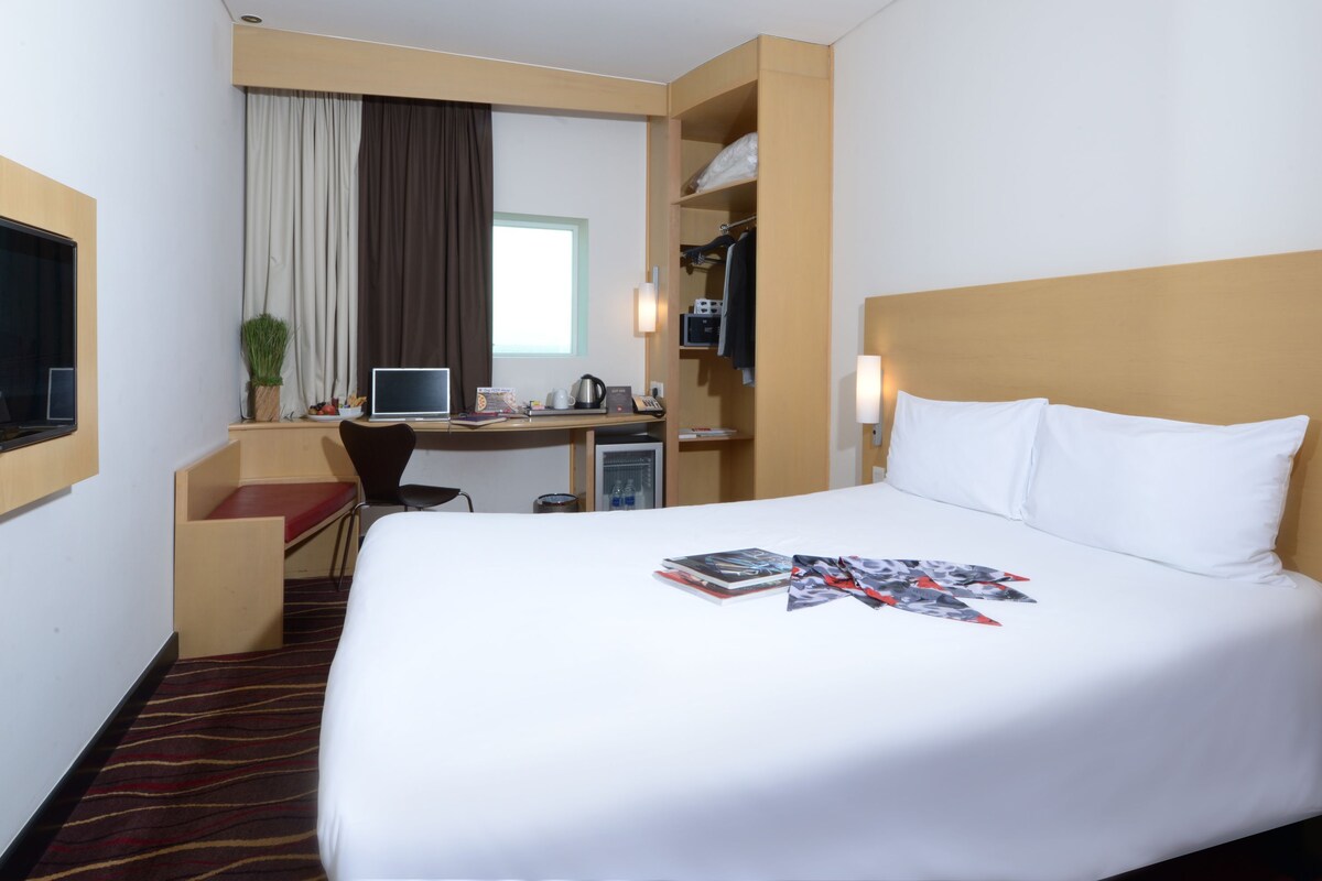 A hotel room with a large bed, a desk with a computer, a chair, and a small table with a vase of flowers.