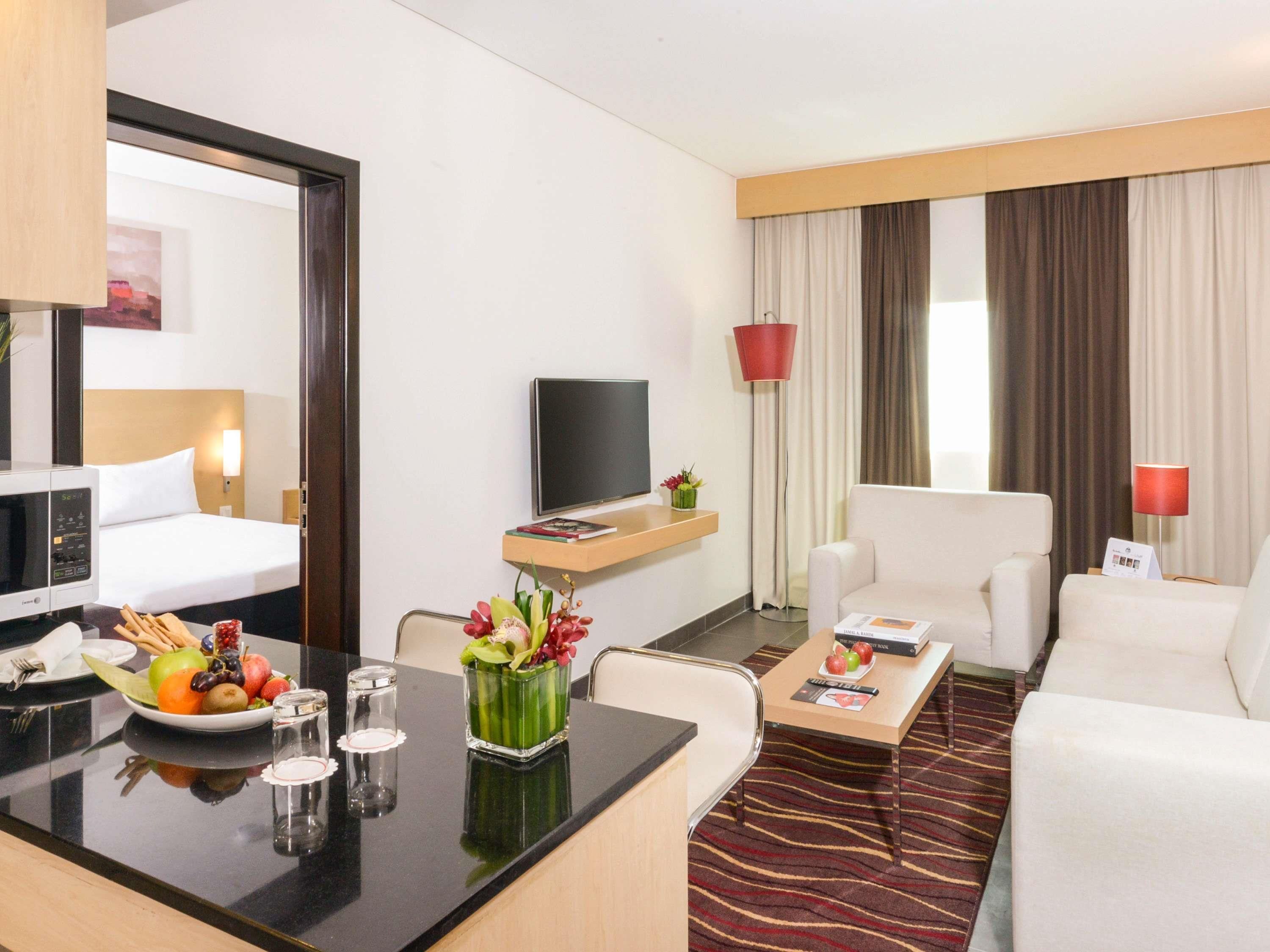 A hotel room with a bed, a desk with a bowl of fruit, a TV, a sofa, and a red lamp.