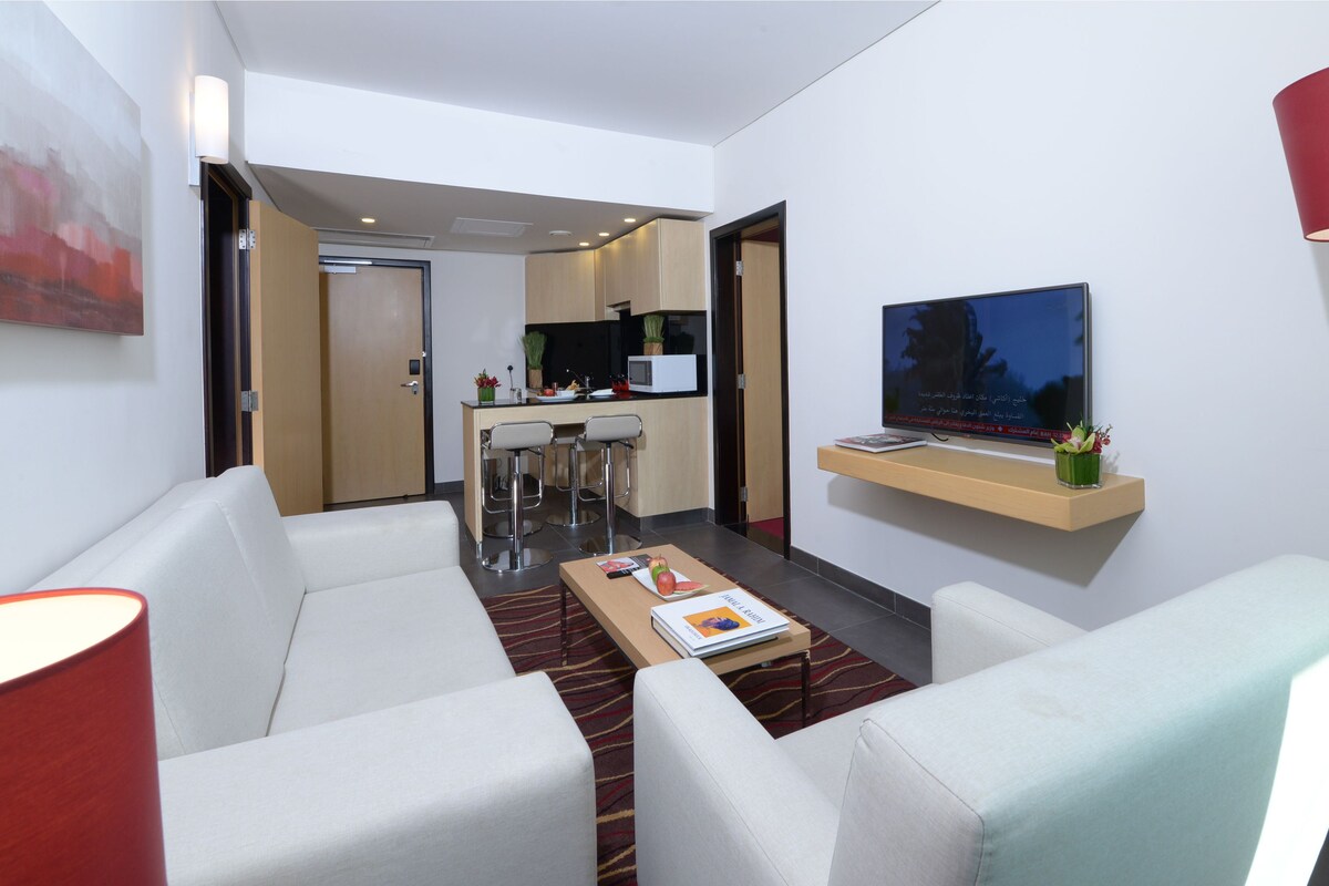A modern living room with a sofa, a coffee table, and a TV mounted on the wall. There is a kitchen area visible in the background.