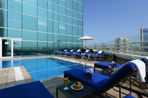 Zona de piscina en la azotea con sillas reclinables azules, una mesa con una bebida y fruta, y barandilla de vidrio con vistas a la ciudad.