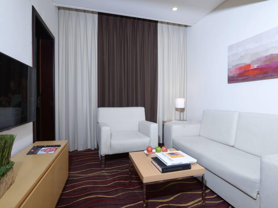 Une chambre d’hôtel équipée d’un téléviseur à écran plat, d’un canapé, d’un fauteuil, d’une petite table avec un livre et des fruits, et d’un tableau accroché au mur.