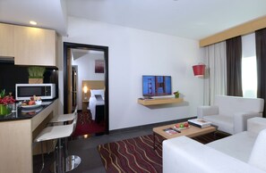 Una habitación de hotel moderna con kitchenette, zona de comedor, sala de estar con sofá y mesa de centro, y un gran ventanal con cortinas.