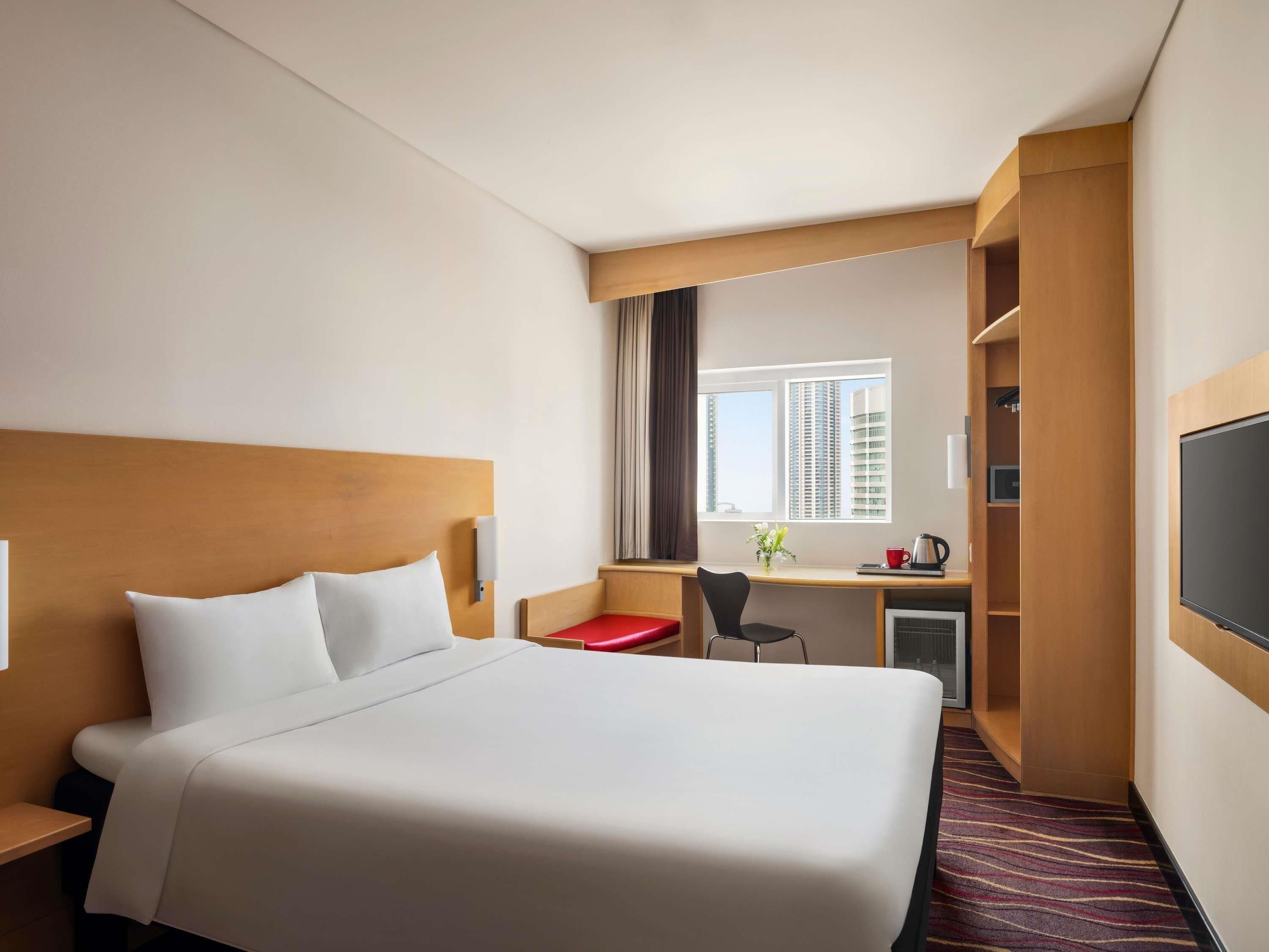 A hotel room with a large bed, a desk with a chair, a TV, and a window with a city view.