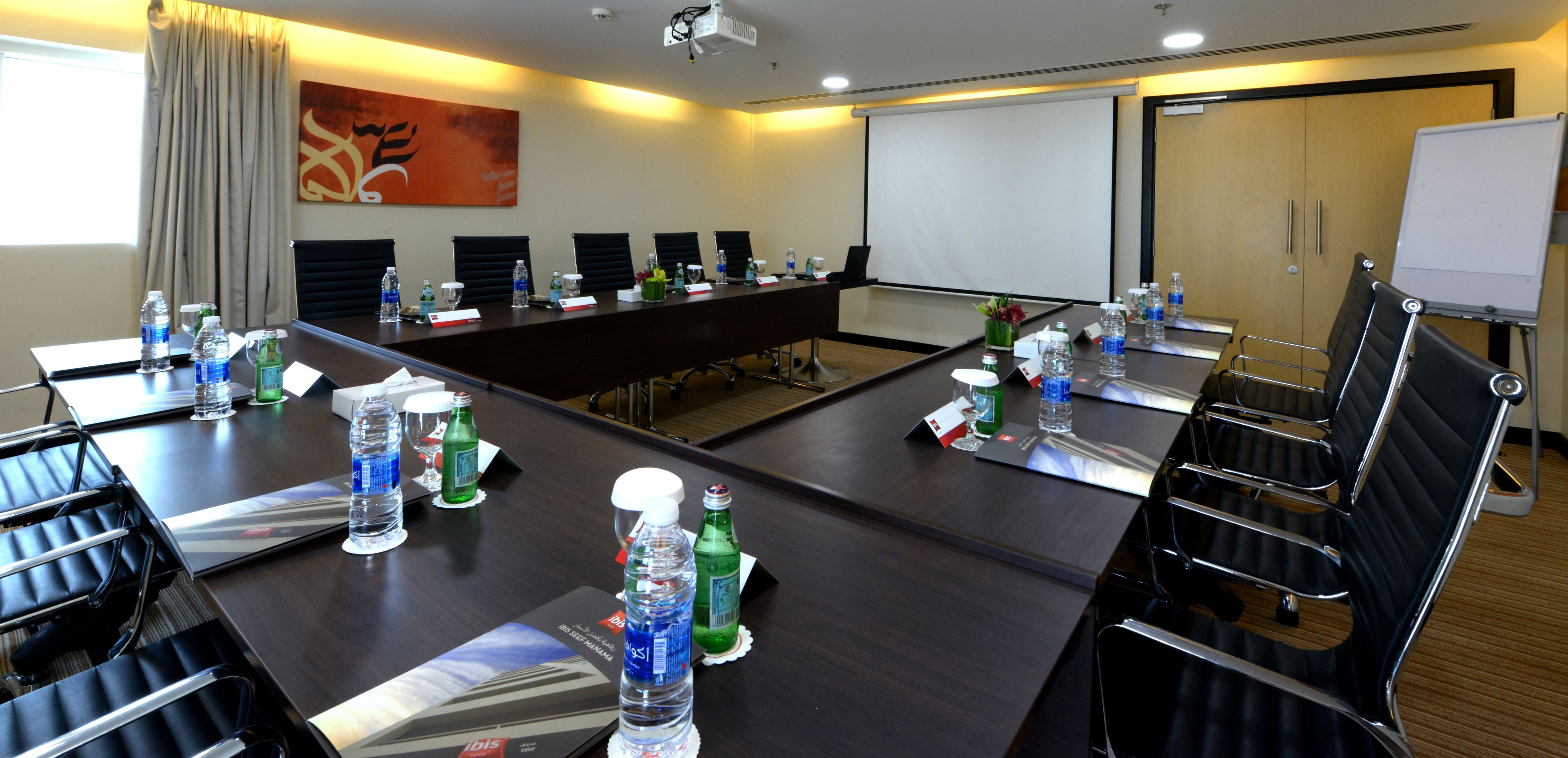 A conference room with a long table, chairs, water bottles, and a projector screen.