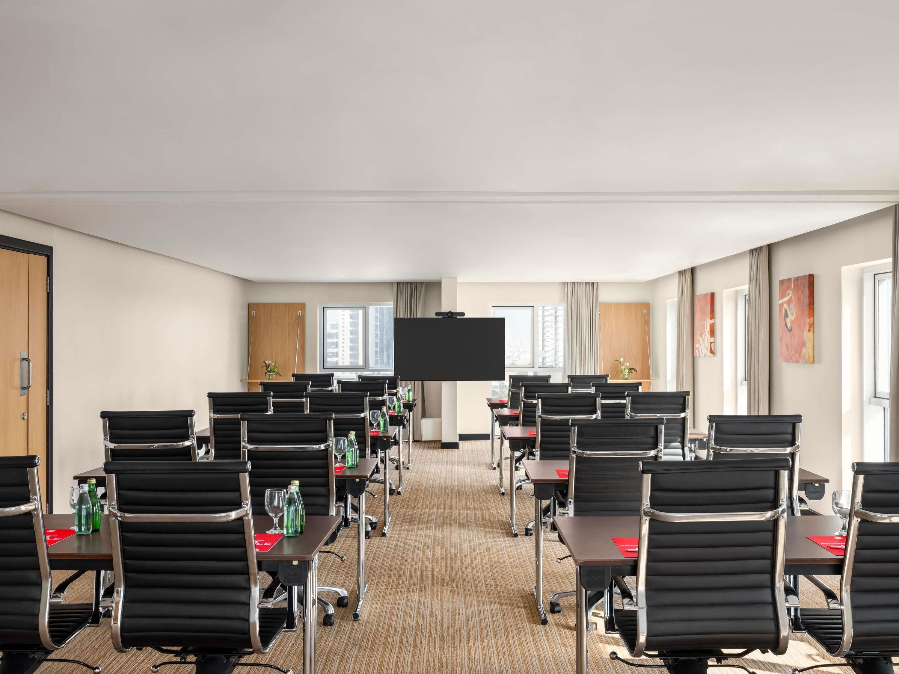 Un salón de conferencias con filas de sillas negras, un televisor de pantalla plana grande y ventanas con cortinas.