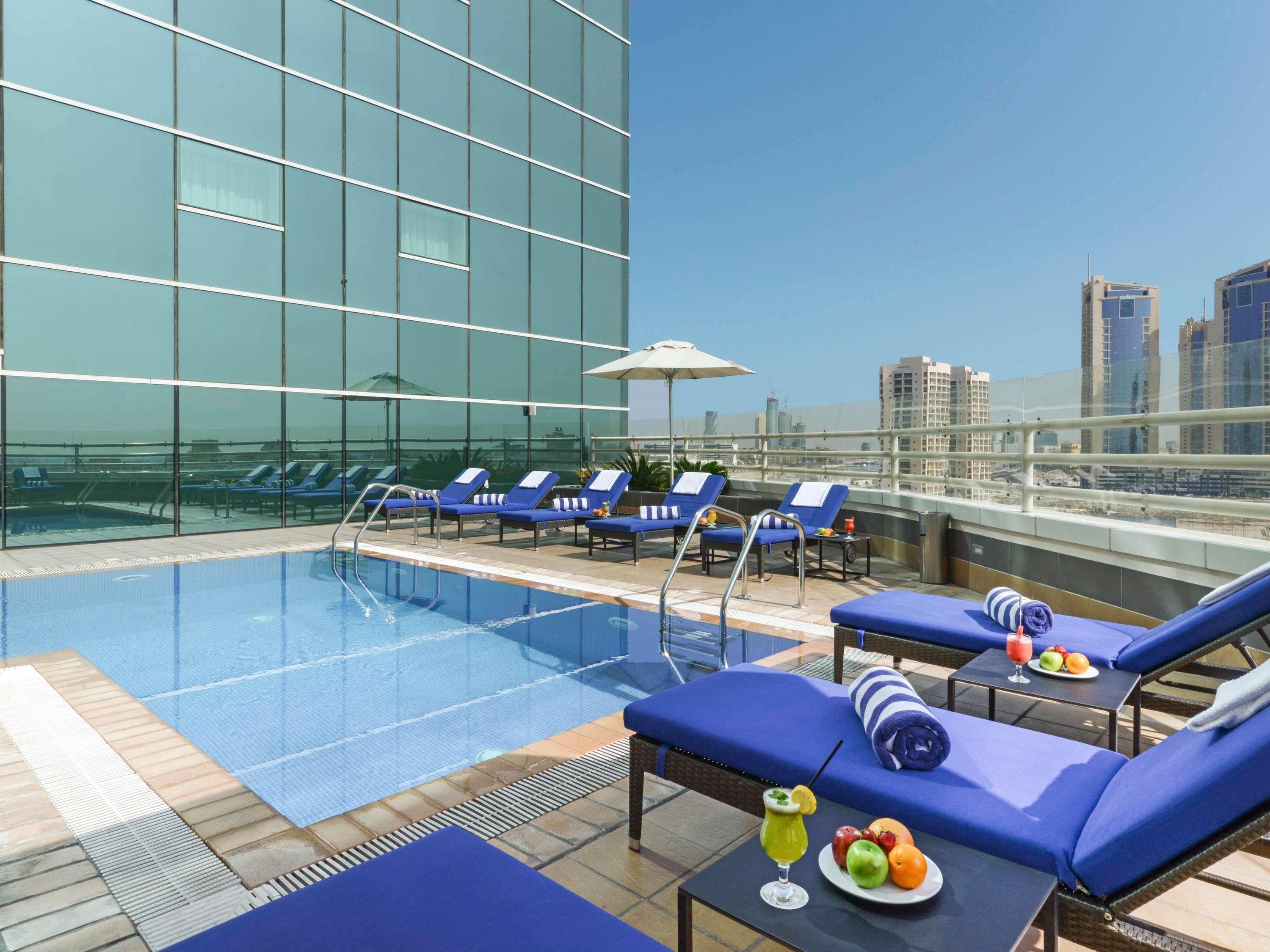 A rooftop pool with lounge chairs, a table with fruit and drinks, and a city view.