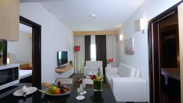 Quarto de hotel moderno com área de refeições, sofá branco, televisão de tela plana e mesa de jantar posta com frutas e copos.