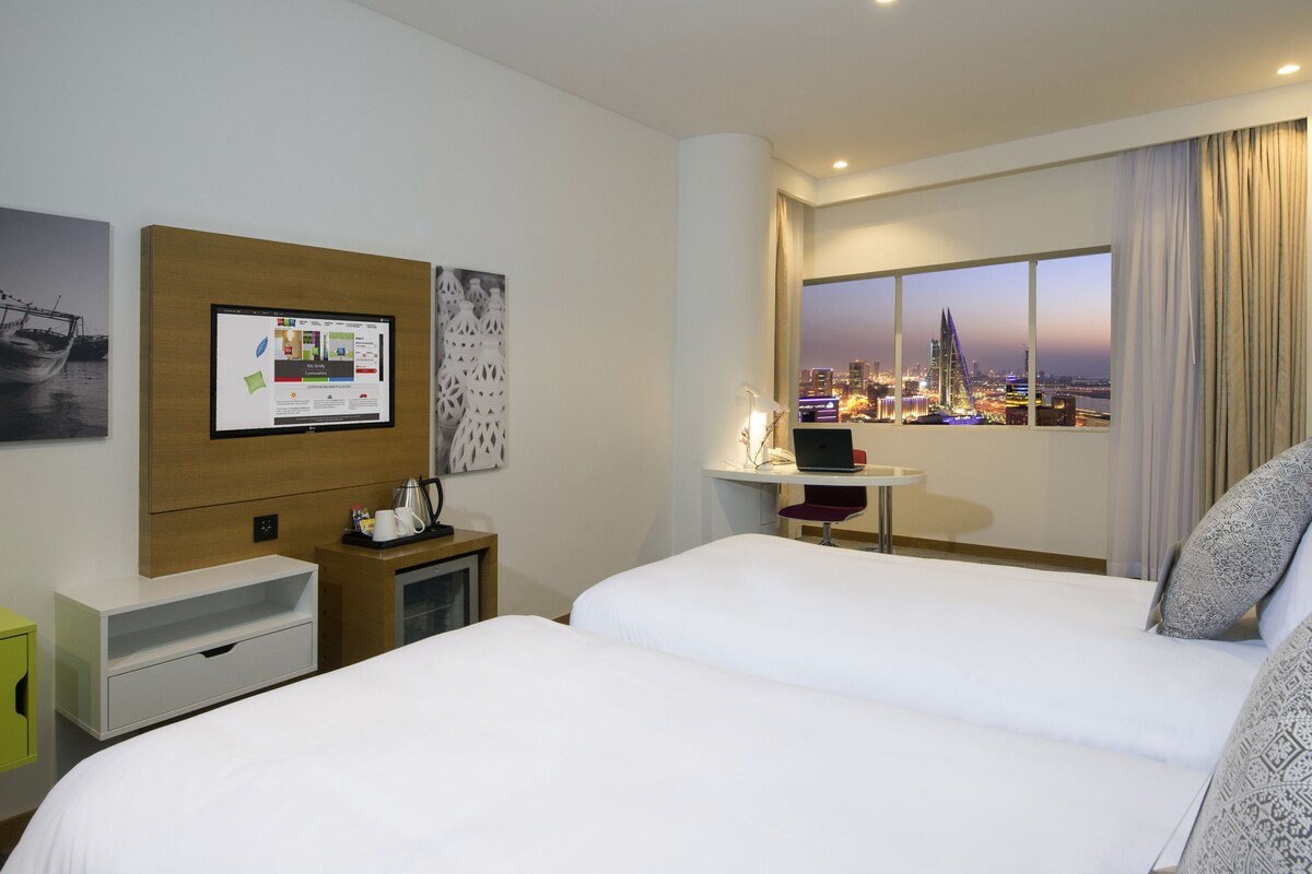A hotel room with a large bed, a desk with a laptop, a TV, and a city view.