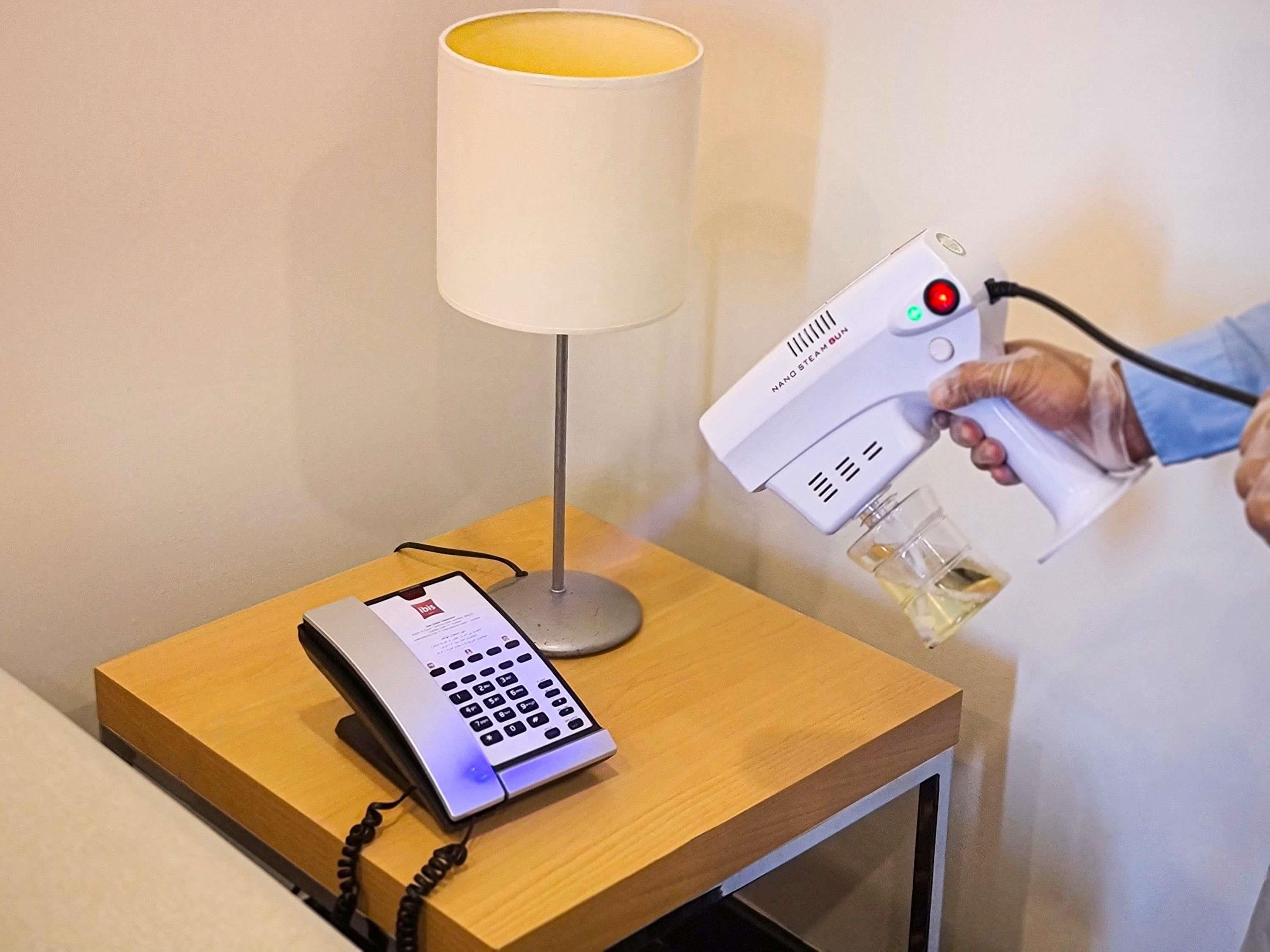 Une chambre d’hôtel avec une table de chevet, une lampe, un téléphone et une personne utilisant un appareil électronique.