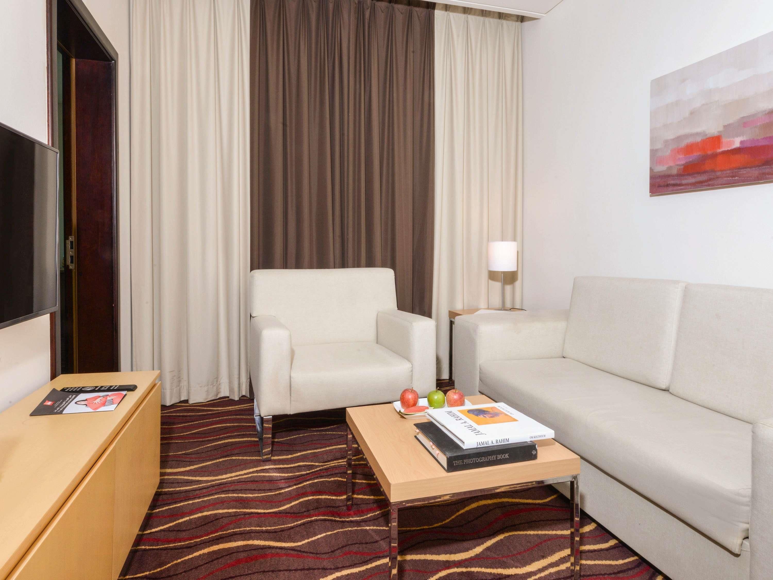 A hotel room with a sofa, armchair, and a small table with a book and fruit.