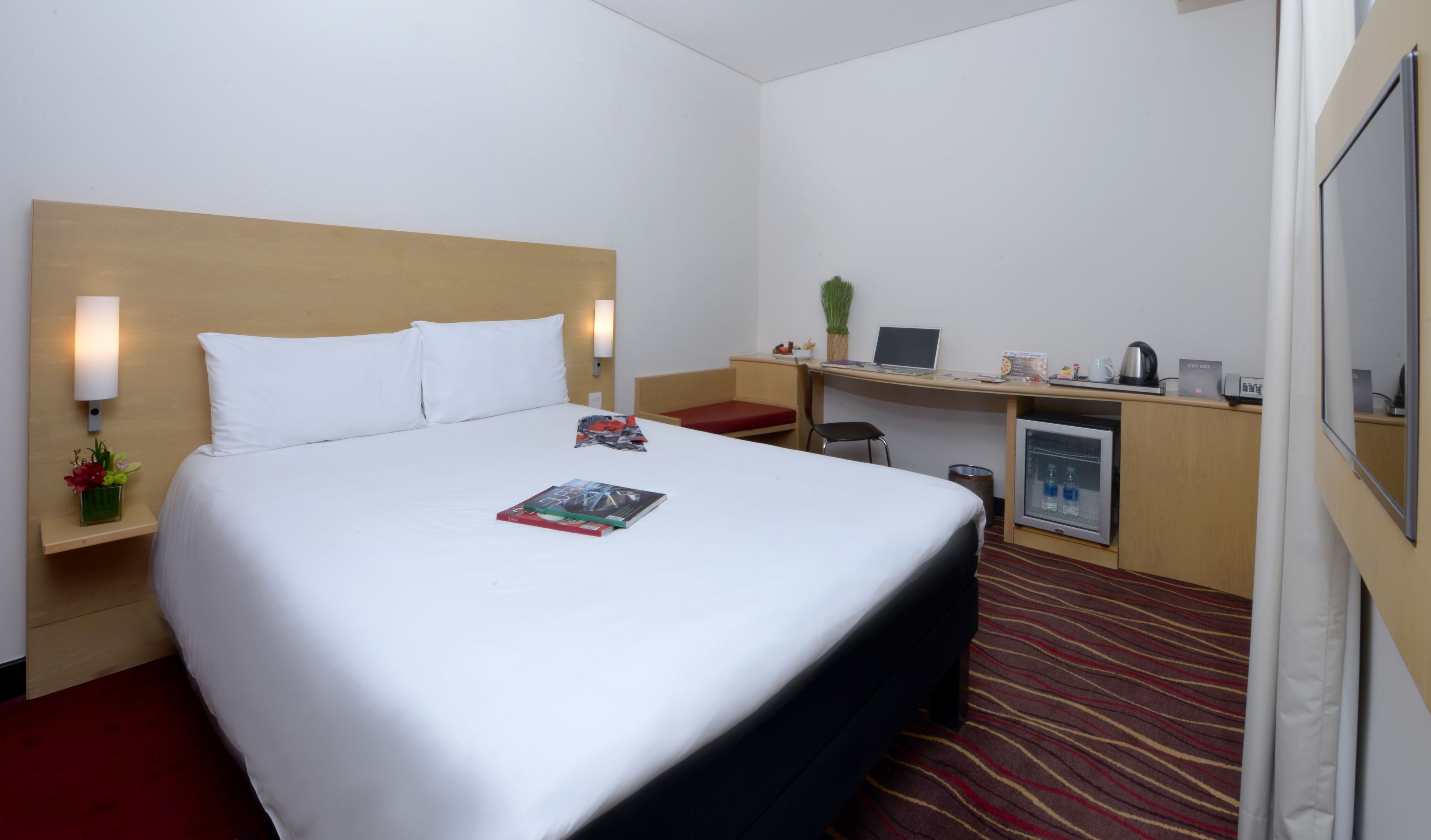 A hotel room with a bed, a desk, a chair, a refrigerator, and a small table with a plant.