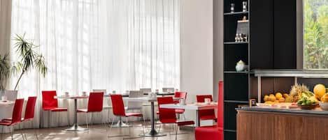 Un comedor moderno con sillas rojas y mesas blancas, una barra negra con frutas y un ventanal con cortinas traslúcidas.