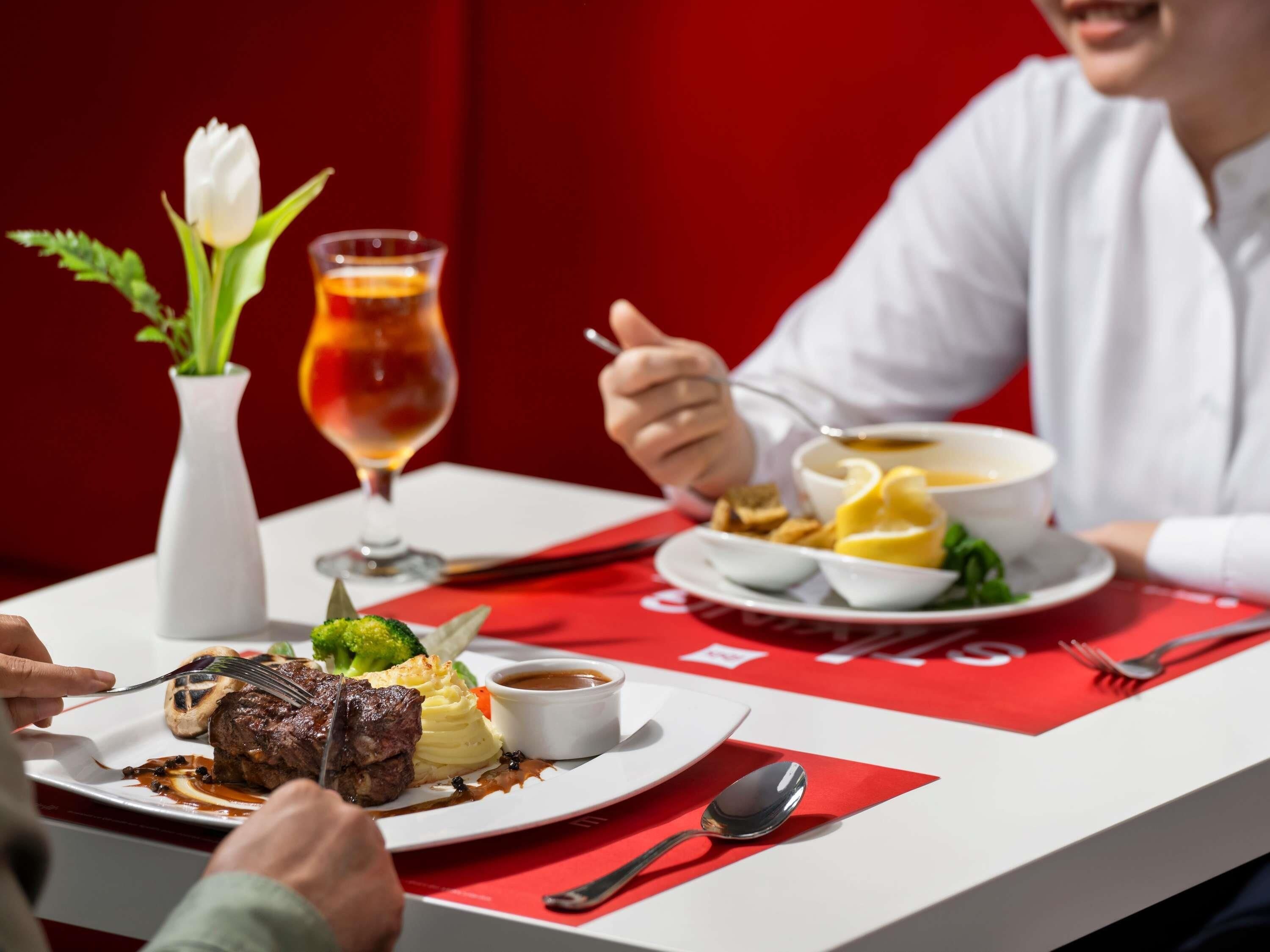 A dining table with a red tablecloth, a white vase with a tulip, a glass of red wine, and a plate of food including steak, mashed potatoes, and vegetables.