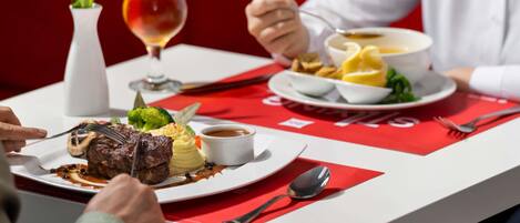 A dining table with a red tablecloth, a white vase with a tulip, a glass of red wine, and a plate of food including steak, mashed potatoes, and vegetables.