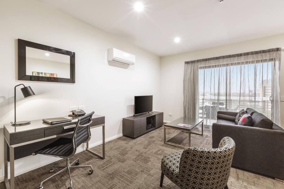 A modern hotel room with a desk, chair, television, sofa, and large window with curtains.