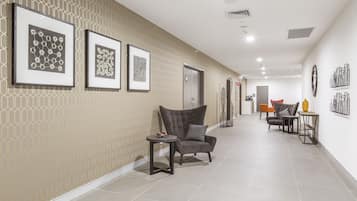 A hallway with framed artwork on the wall, a grey armchair, a small table, and a door in the background.