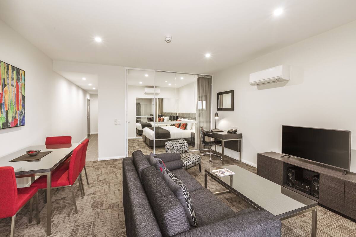A modern living room with a sofa, red chairs, a glass coffee table, a flat-screen TV, and a dining area with a table and chairs.