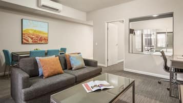 Una habitación de hotel moderna con un sofá gris, una mesa de centro de vidrio y un comedor con una mesa y sillas.