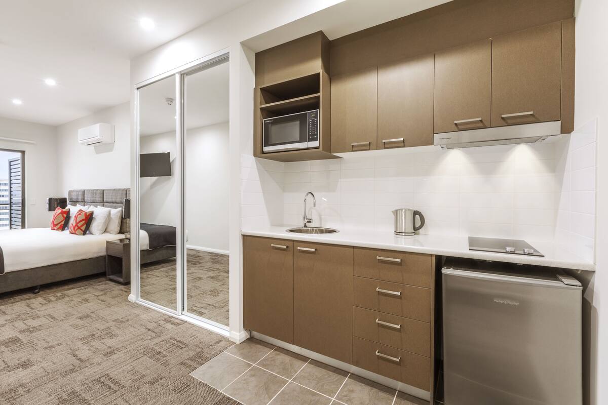 A modern kitchen with a microwave, sink, and refrigerator, adjacent to a bedroom with a bed and nightstand.