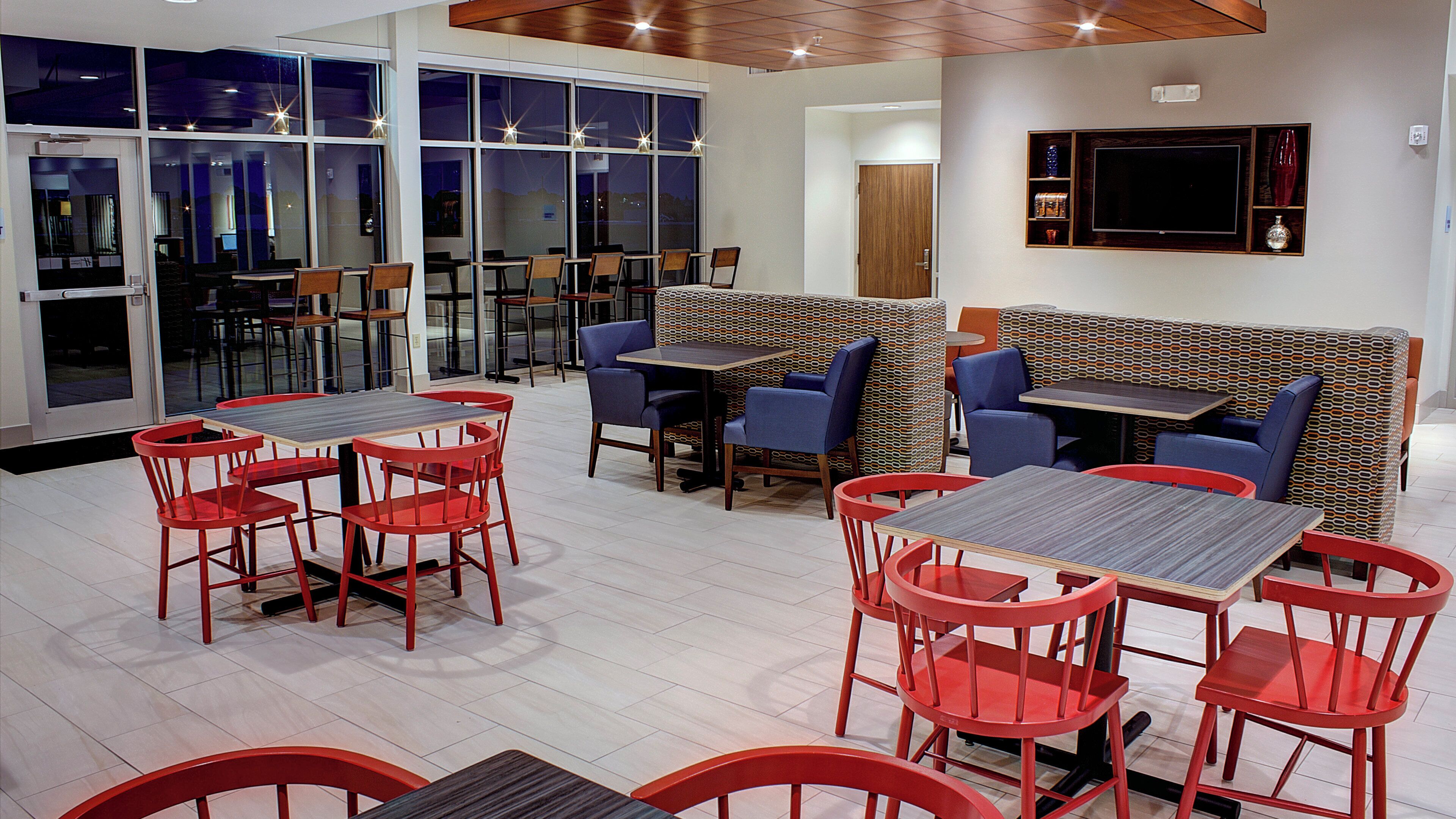 A restaurant with booths, tables, and chairs. There are large windows and a flat-screen TV on the wall.