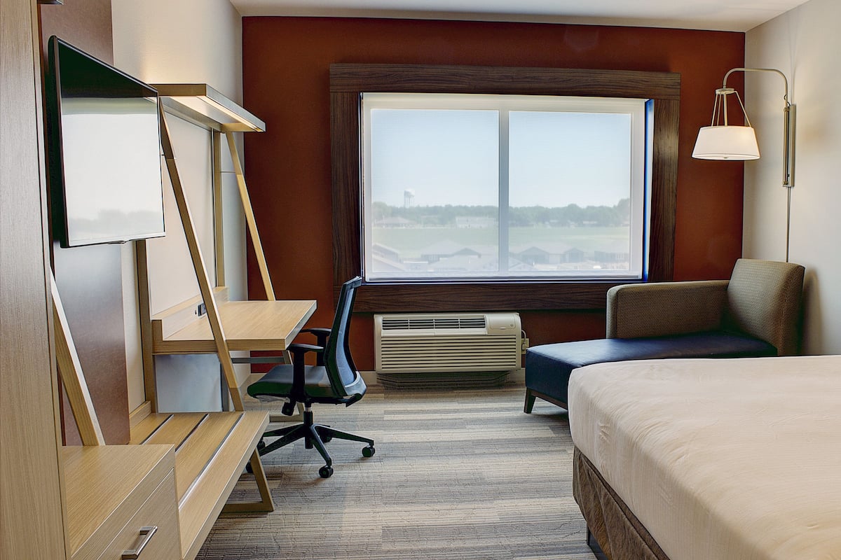 A hotel room with a bed, a desk with a chair, a window with a view, and a wall-mounted lamp.