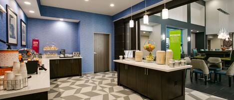 A modern hotel lobby with a geometric patterned floor, a reception desk with a coffee station, and a seating area with chairs and tables.